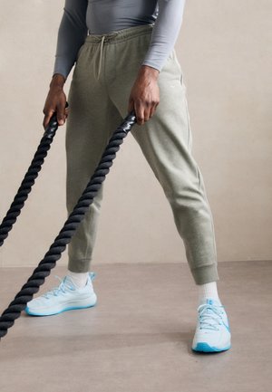 Persona che indossa una maglia grigia a maniche lunghe, pantaloni della tuta grigio chiaro e scarpe da ginnastica blu mentre tiene delle corde da battaglia nere in un ambiente per allenamenti al chiuso.