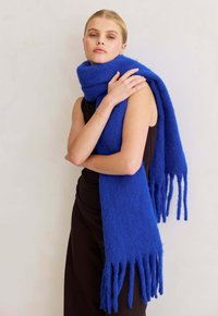 Vrouw in een mouwloos zwart jurkje met een dikke, lange blauwe sjaal om haar nek en schouders, tegen een effen achtergrond.