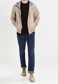 Beige zip-jacka med en blå fodrad huva, mörk tröja, mörka uppvikta jeans och ljusa stövlar. Slät textur och avslappnad passform.