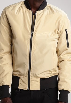 Mann trägt beige Bomberjacke mit schwarzem Kragen, Bündchen, Bund und Reißverschlusstasche am linken Ärmel, Frontreißverschluss geschlossen.