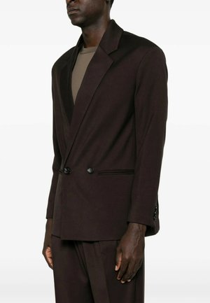 Uomo che indossa un blazer doppiopetto marrone scuro e pantaloni abbinati sopra una camicia color taupe, in piedi davanti a uno sfondo chiaro e uniforme.