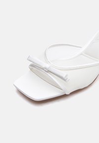 Sandale blanche à bout ouvert avec une fine bride et un petit nœud décoratif sur le devant, sur un fond uni.