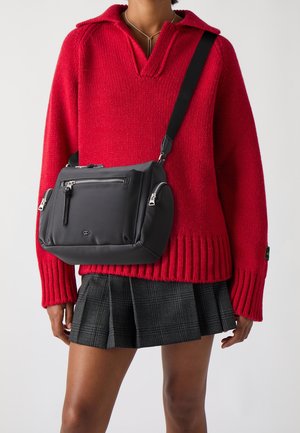 Zwarte nylon crossbodytas met zilveren ritsen en dubbele zijzakken, gedragen over een rode gebreide trui en geruite rok. Voorzien van een afneembare riem.