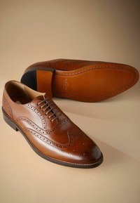 Ruske nahkasaappaat broguedetaililla, kappeetolla ja nauhatuella. Sujuva pinta, jossa on kontrastiväri musta korko ja ruskea pohja.
