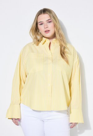 Camicia gialla a righe con maniche lunghe e polsini svasati, dotata di tasca frontale. Abbinata a pantaloni bianchi.
