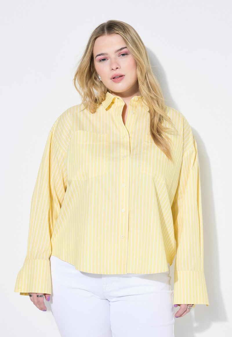 Gelb gestreifte Button-Down-Bluse mit langen Ärmeln und ausgestelltenden Manschetten, ausgestattet mit einer Brusttasche. Kombiniert mit weißen Hosen.