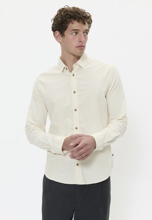 Lys beige skjorte med knapper lavet af bomuld, med lange ærmer, krave og brune knapper. Bæres med mørke bukser.