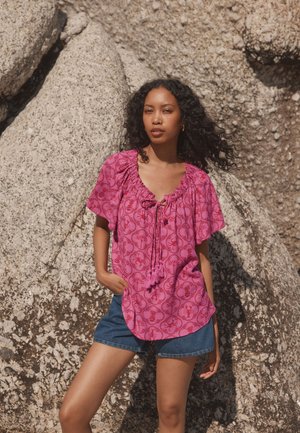 Blusa rosa con motivo, maniche corte e larghe e scollo con coulisse, abbinata a pantaloncini di jeans, su sfondo roccioso.