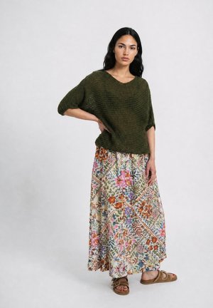 Frau trägt einen grünen locker gestrickten Pullover, floralen Maxirock und braune Sandalen und steht mit einer Hand in der Hüfte vor einem schlichten Hintergrund.