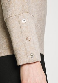 Beiger Strickpullover-Bündchen mit drei kleinen, irisierenden Knöpfen, weicher Textur und subtil gemustertem Melange-Design, kombiniert mit einem schwarzen Kleidungsstück.