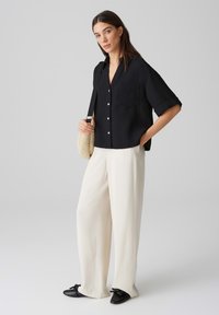 Maglietta nera a maniche corte con texture e tasca, abbinata a pantaloni bianchi a gamba larga e scarpe nere con dettagli a fiocco. Sfondo chiaro.