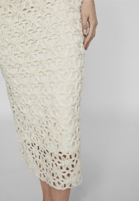 Hvit blonde midi-skjørt med blomsterdesign og lagdelte utskjæringer. Stoffet har en teksturert overflate og en litt gjennomsiktig utseende.