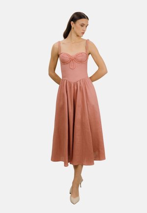 Kvinne med mørkt hår iført en ermeløs midi-kjole i støvete rosalilla med flettede stropper og beige spisse hæler, stående med hendene bak ryggen.