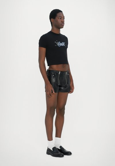Tricou scurt negru cu text brodat, purtat alături de pantaloni scurți din piele neagră cu fermoare, mocasini negri și șosete albe până la gleznă.