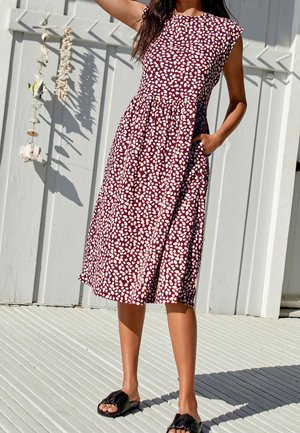 Robe bordeaux à motif de pois blancs, manches courtes, col rond, taille cintrée et poches. Portez avec des sandales plates noires sur une surface texturée.