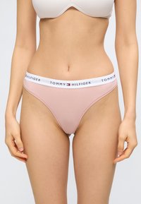 Γυναίκα που φοράει ανοιχτό ροζ μπικίνι εσώρουχο με λευκή ζώνη μέσης που φέρει το λογότυπο Tommy Hilfiger, στέκεται μπροστά σε απλό φόντο.