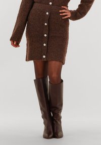 Bruine gebreide jurk met een aansluitende silhouet, voorzien van zeven grote witte knopen aan de voorkant. Bruine hoge leren laarzen maken de look compleet.