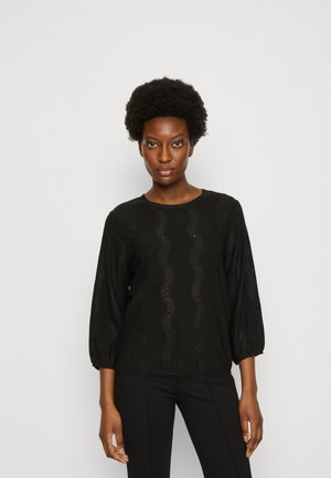 Blouse noire à manches longues avec un motif en dentelle ondulée, col rond et manches froncées. Associée à un pantalon noir ajusté.