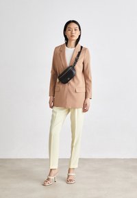 Blazer marrone chiaro con tasche a patta, top bianco, pantaloni gialli e borsa nera a tracolla. La modella indossa sandali con tacco chiari.