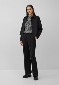Vrouw die met handen in de zakken staat, gekleed in een zwarte cropped jas, grijs panterprint trui, zwarte broek en zwarte chunky schoenen.