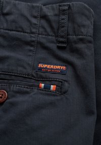 Marineblåt stof med et "SUPERDRY®" mærkat i orange og hvid tekst samt en lille broderet detalje med orange, blå og hvide striber.