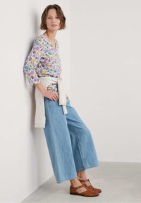 Blusa con motivo floreale, con scollatura a V e maniche a tre quarti, abbinata a jeans blu a gamba larga e ballerine intrecciate marroni. Maglione bianco in maglia legato in vita.