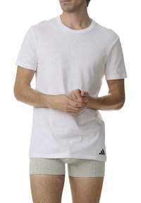 Biała bawełniana koszulka z krótkim rękawem, okrągłym dekoltem i czarnym logo Adidas na lewej stronie. Pod spodem założone są szare majtki typu bokserki.