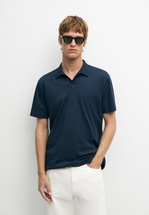 Junger Mann mit schwarzen Sonnenbrillen, einem dunkelblauen kurzärmeligen Poloshirt und weißen Hosen, steht mit einer Hand in der Tasche vor einem einfarbigen Hintergrund.