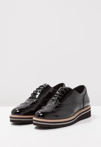 Black patent leather shoes featuring brogue detailing, round toes, and a contrasting white and black sole. Laced design with smooth finish.