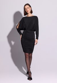Czarna sukienka z długimi rękawami, zdobionym tiulem na biuście i marszczonym dolnym fragmentem. Gładka tkanina o delikatnym połysku; modelka nosi przezroczyste rajstopy i czarne szpilki.