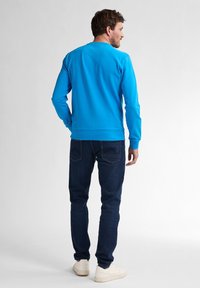 Camicia blu a maniche lunghe con scollatura rotonda e polsini a coste, abbinata a jeans blu scuro e sneaker di colore chiaro.