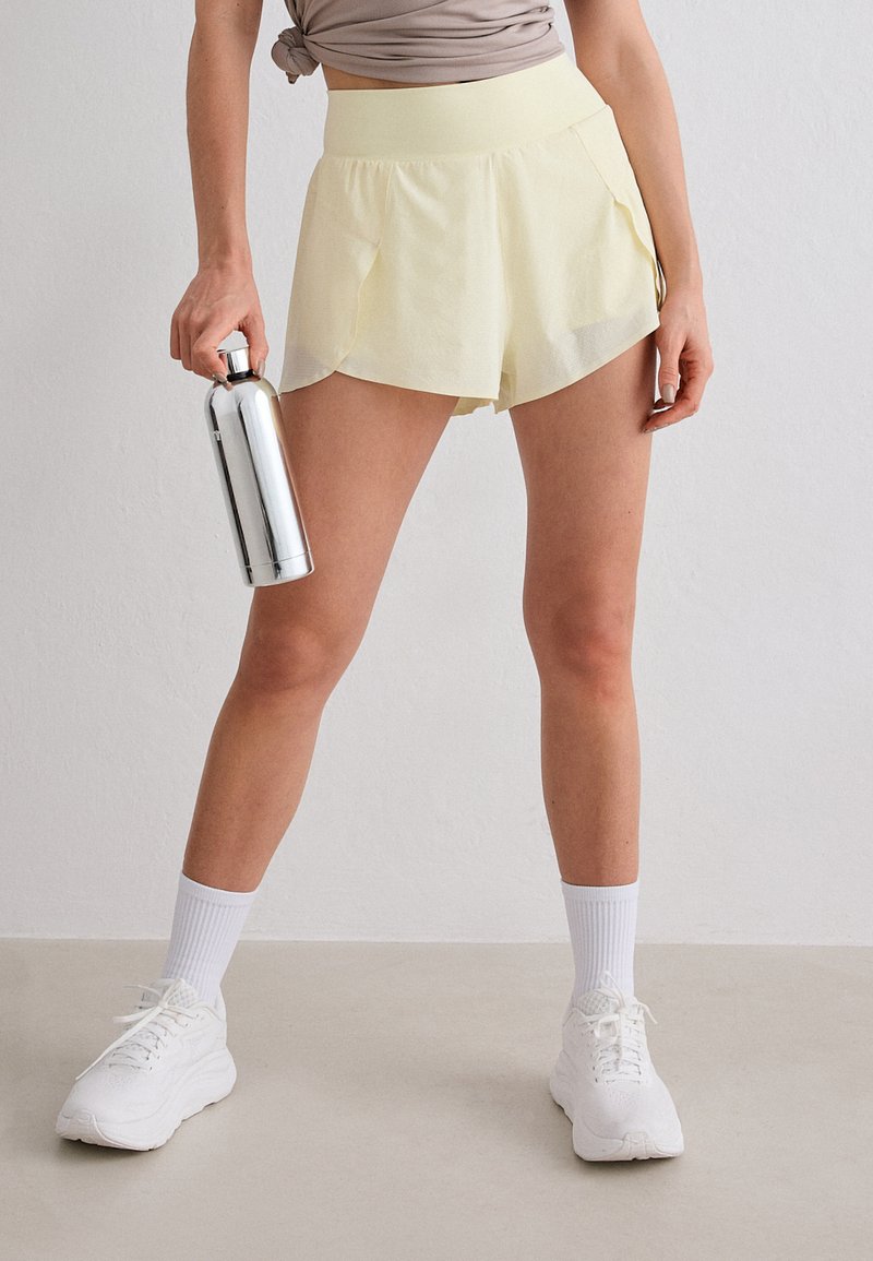 Femme portant un short de sport jaune pâle et des baskets blanches, tenant une bouteille d'eau argentée devant un mur gris uni.