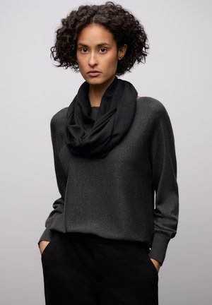 Zwarte sjaal met een lus, gedrapeerd over een donkergrijze, getextureerde trui met brede, ontspannen mouwen. De outfit is gecombineerd met zwarte broeken.