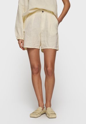 Kobieta nosząca kremowe lniane szorty i dopasowaną bluzkę z długim rękawem oraz kremowe wsuwane mokasyny, stoi z jedną ręką w kieszeni.