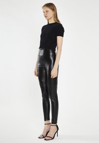 Leggings neri in pelle sintetica con vita alta, abbinati a un top corto fluffy nero a maniche corte. La modella indossa tacchi neri con le cinghie.
