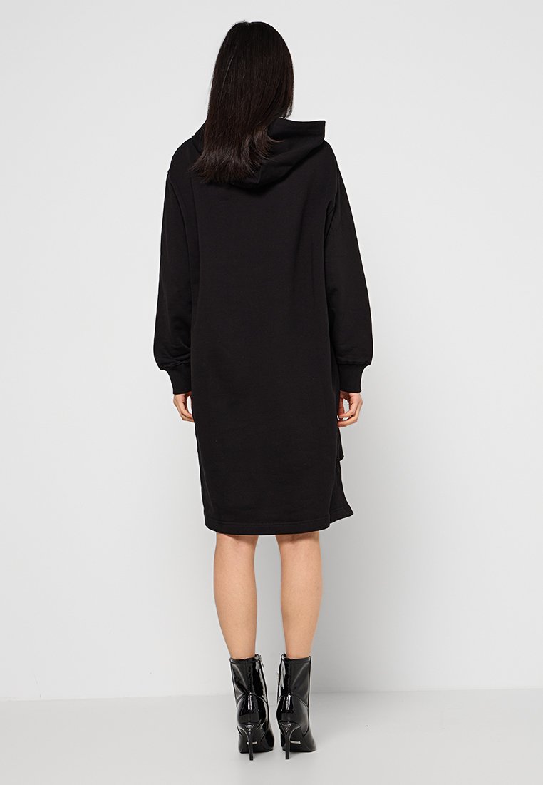 Vrouw die met haar rug naar de camera staat en een zwart gewatteerd sweatshirt-dress met capuchon draagt, samen met zwarte laarzen met hoge hakken van glanzend leer.