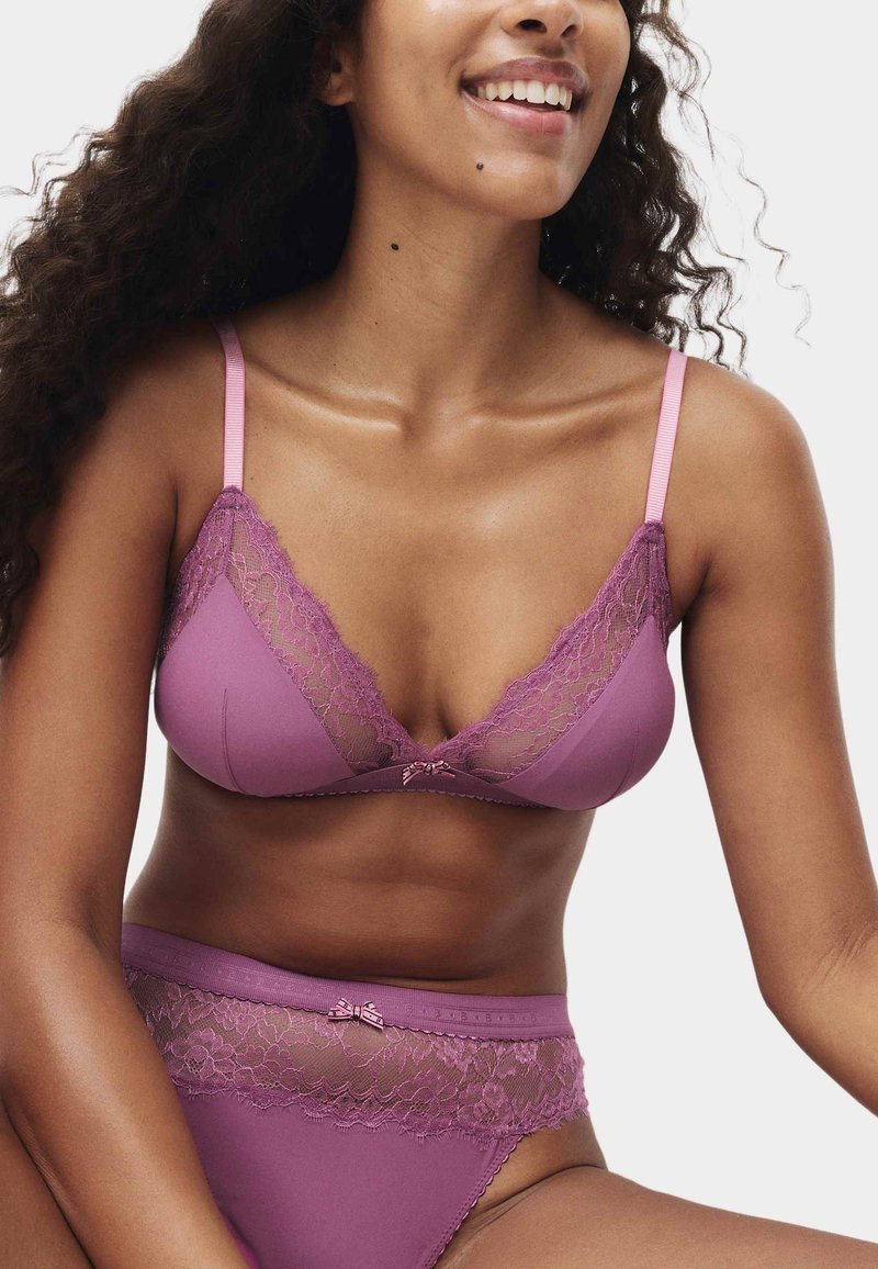 Kobieta w fioletowym biustonoszu z koronkową lamówką i pasujących majtkach z wysokim stanem, uśmiechająca się z kręconymi włosami na białym tle.