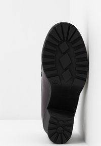 Semelle d'une botte noire avec des motifs de crampons profonds, trois formes de losange au centre, positionnée contre une surface d'angle blanche.
