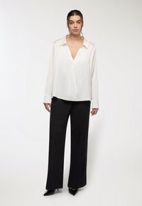 Biała bluzka z długim rękawem i dekoltem w kształcie V; zestawiona z czarnymi spodniami o szerokich nogawkach. Błyszczące, czarne szpilki podkreślają stylizację. Gładka tekstura materiału.