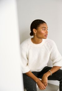 Donna con capelli intrecciati, che indossa un maglione bianco a maglia e pantaloni neri con dettagli argentati, è seduta e guarda verso destra, appoggiata su una parete chiara e semplice.