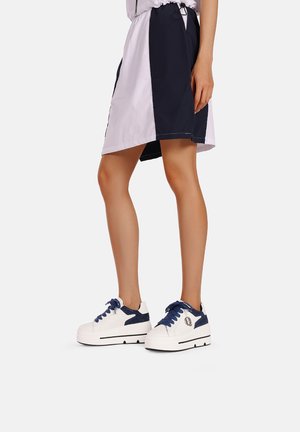 Sneakers bianchi con accenti e lacci blu navy, indossati con una gonna al ginocchio a blocchi di colore in viola e blu navy. Texture liscia, design casual.