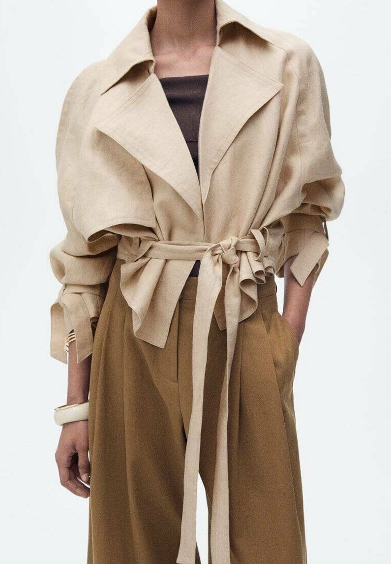 Veste beige courte avec des revers larges, des manches fronc ées et une taille à nouer. Portée sur un haut marron foncé, associée à un pantalon beige taille haute.