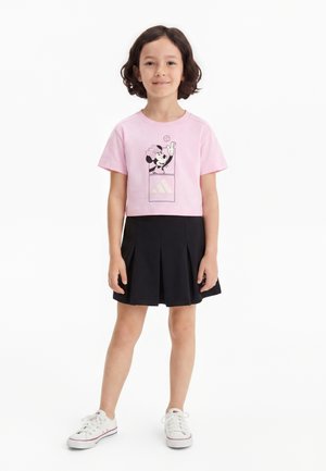 Enfant aux cheveux courts bouclés portant un t-shirt rose à motif Minnie Mouse, une jupe plissée noire et des baskets blanches, debout sur fond blanc.