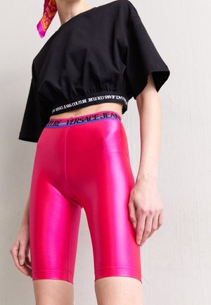 Persoon draagt glanzende roze high-waisted biker shorts met Versace Jeans tailleband en een losse zwarte cropped top met tekstdetail.