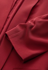 Material burgundy cu o suprafață netedă și texturată. Prezintă o mânecă pliată, evidențiind un design croit și cusături curate de-a lungul marginilor.