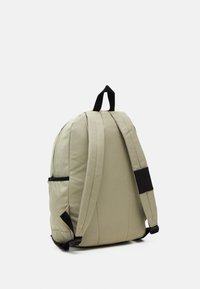 Mochila beige con lados acolchados, correas ajustables y detalles en negro. Cuenta con un asa en la parte superior y un bolsillo lateral. Textura de tela suave.