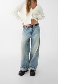 Bílý žebrovaný cardigan, zkrácená délka, nošený přes bílý top. Svetle modré široké džíny s vybledlými místy, doplněné černými platformovými botami a opaskem se cvočky.