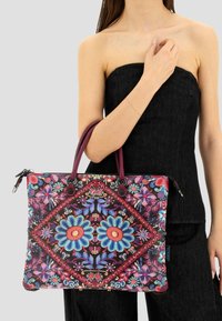 Donna in un abito nero senza spalline che tiene una grande borsa a tracolla con ricami floreali colorati e manici bordeaux.