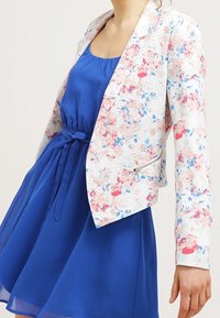 Floral blazer with a white base featuring pink and blue flowers, paired with a blue dress. Blazer has a structured design and zip details.