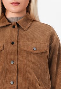 Gros plan d'une personne portant une veste en velours côtelé marron avec des boutons-pression noirs et un col, par-dessus un pull à col roulé côtelé gris.